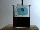 Prusion Blue Film hanging from rod into top half of beaker whose bottom half is filled with dark liquid